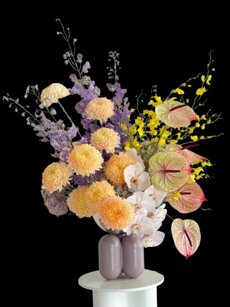 A vase of dahlias, anthuriums and larkspur on a pale plinth