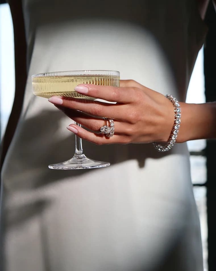 A hand holding a coupe, a cocktail ring and a tennis bracelet