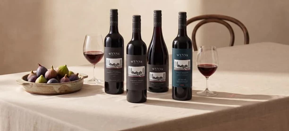 A row of red wine bottles on a linen table beside figs and a poured glass